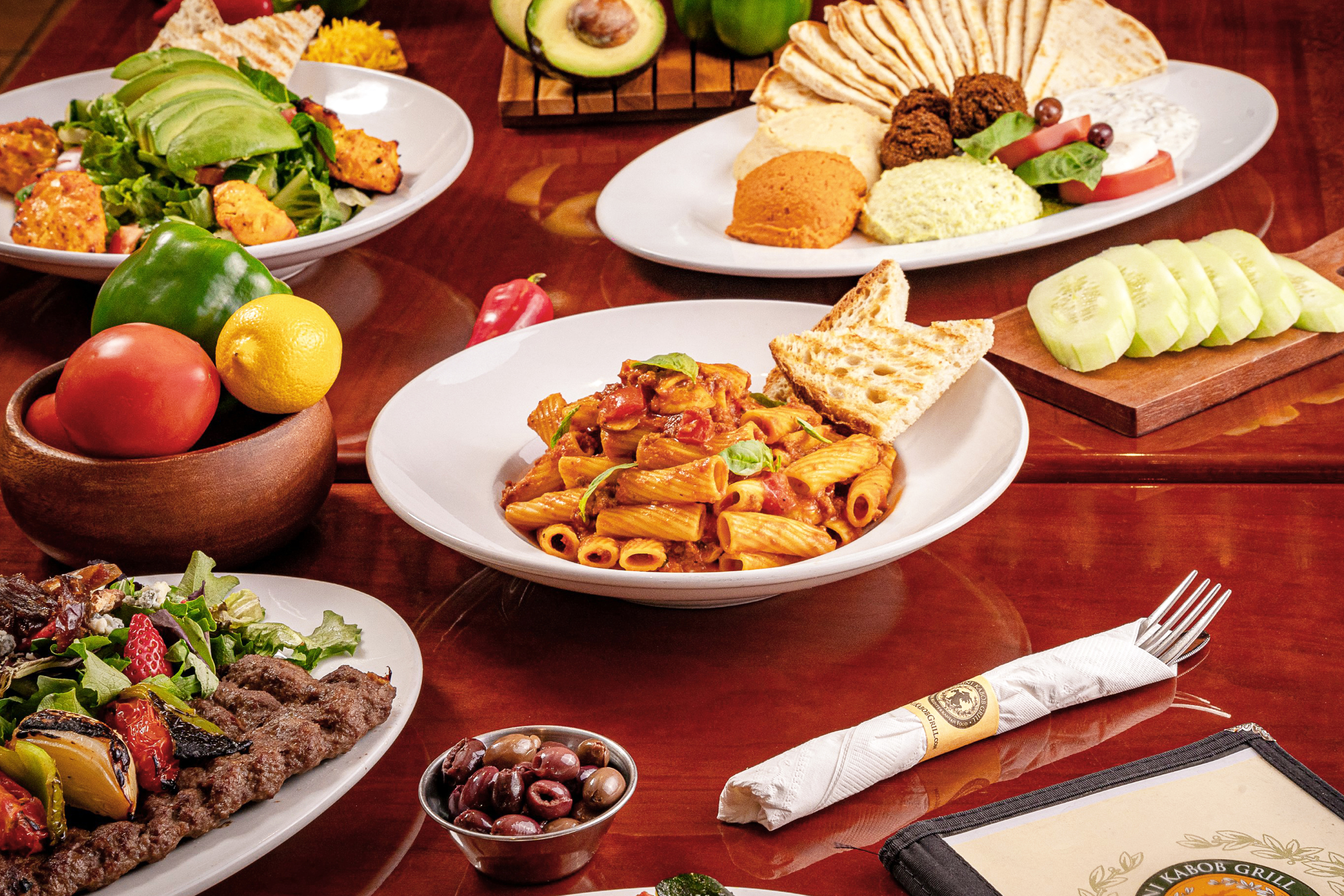 Panini Kabob Grill table spread featuring rigatoni pasta, grilled beef koobideh kabob with salad, chicken avocado salad, and a mezze platter with hummus, falafel, and pita